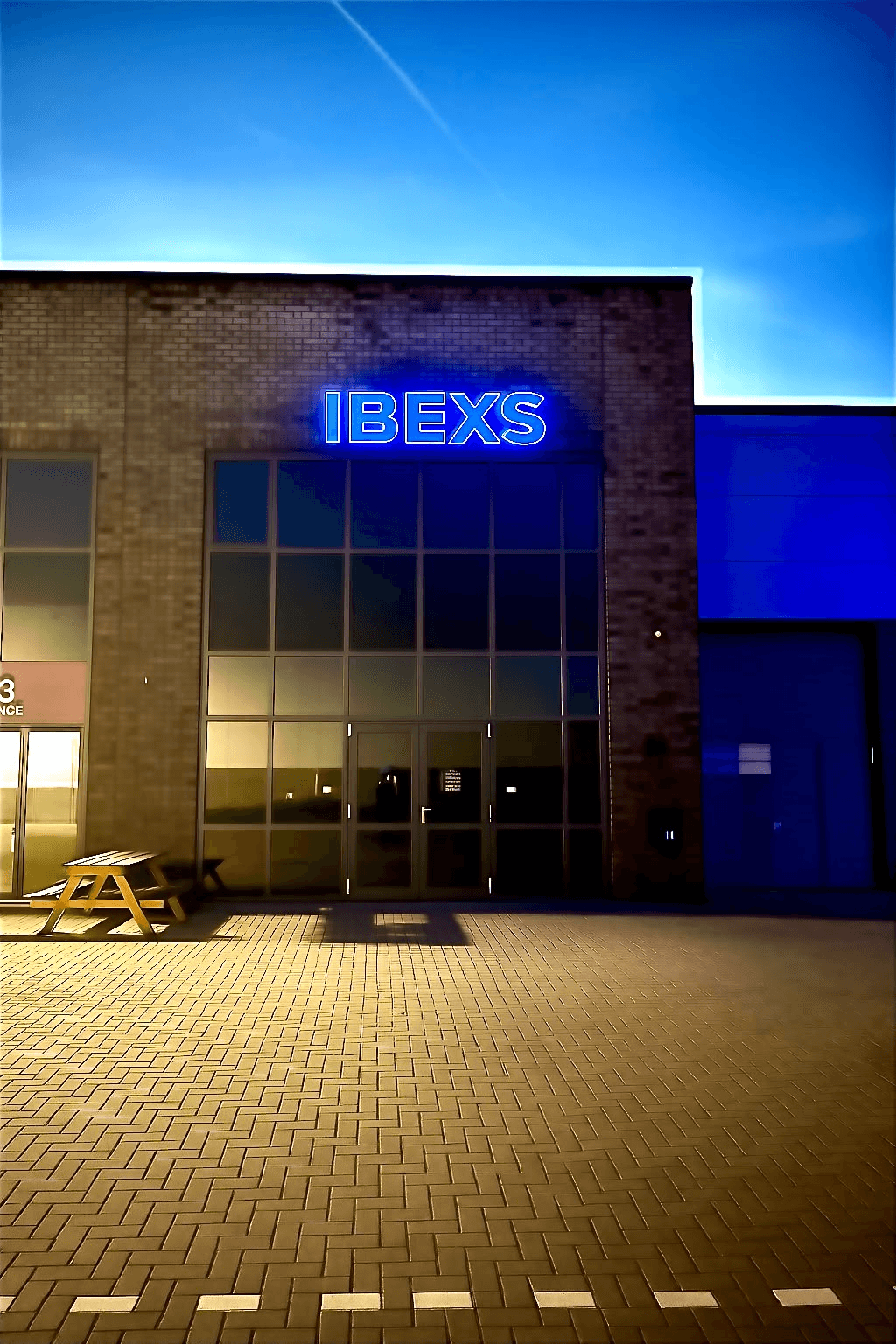 Bakstenen gevel met een blauw verlicht IBEXS-logo boven een glazen entree.
