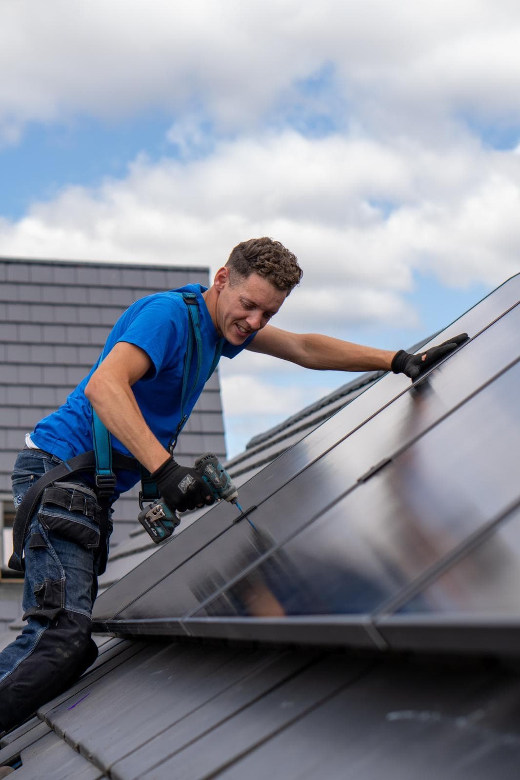 Een man met veiligheidsharnas werkt met een accuboormachine aan zonnepanelen op een schuin dak.